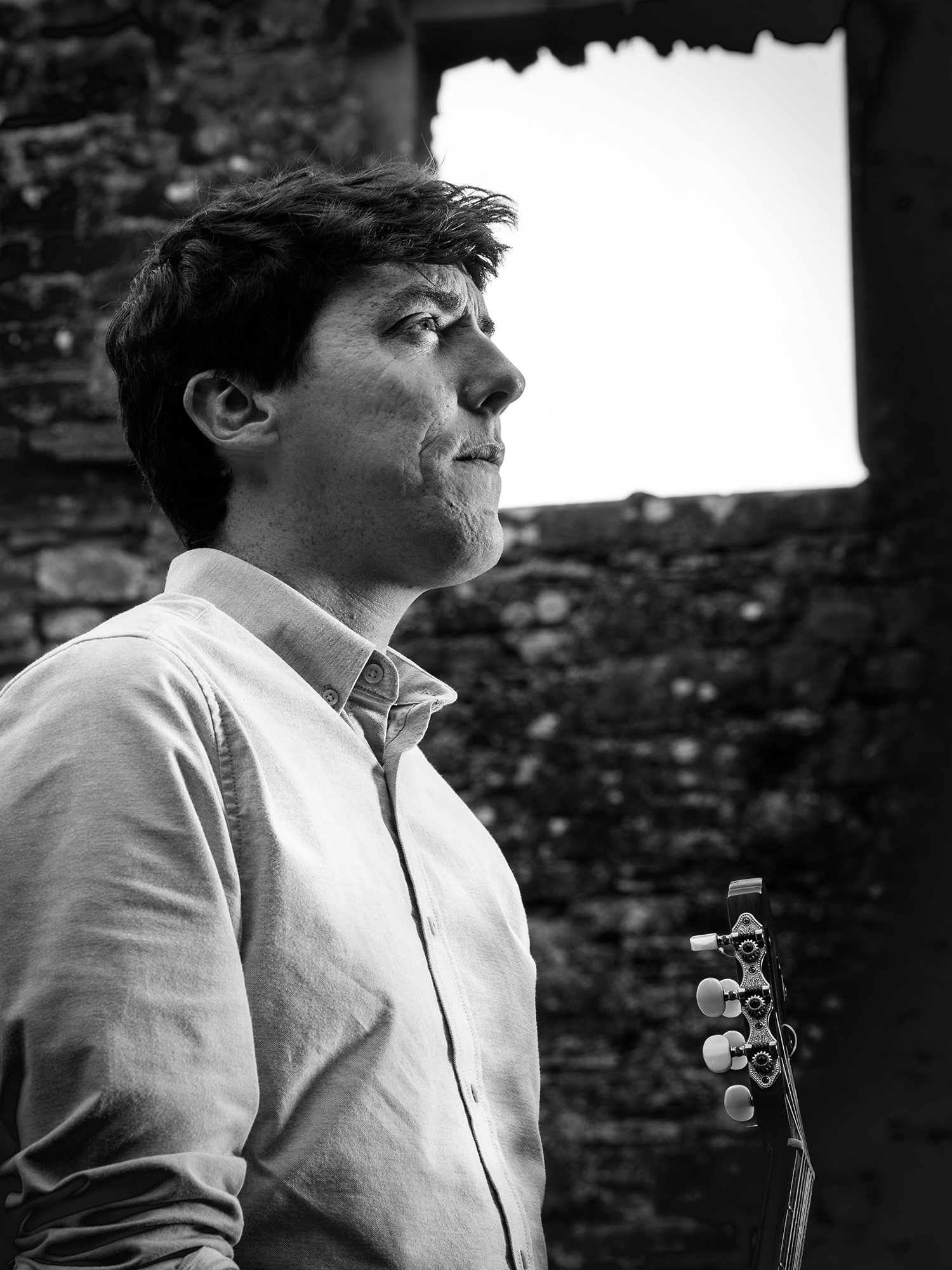 Black and white portrait of Oscar Montague holding a guitar, standing in front of weathered stone ruins and gazing thoughtfully into the distance.
