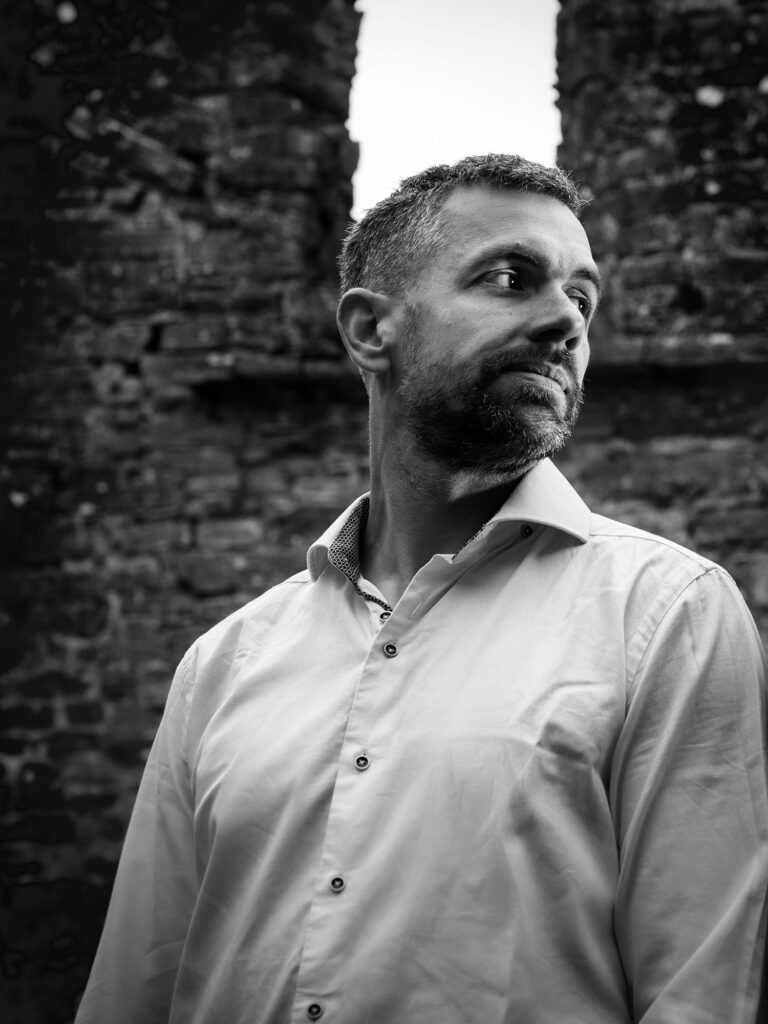 Black and white portrait of Santiago Kodela standing in front of historic stone ruins, looking off to the side in a reflective pose.